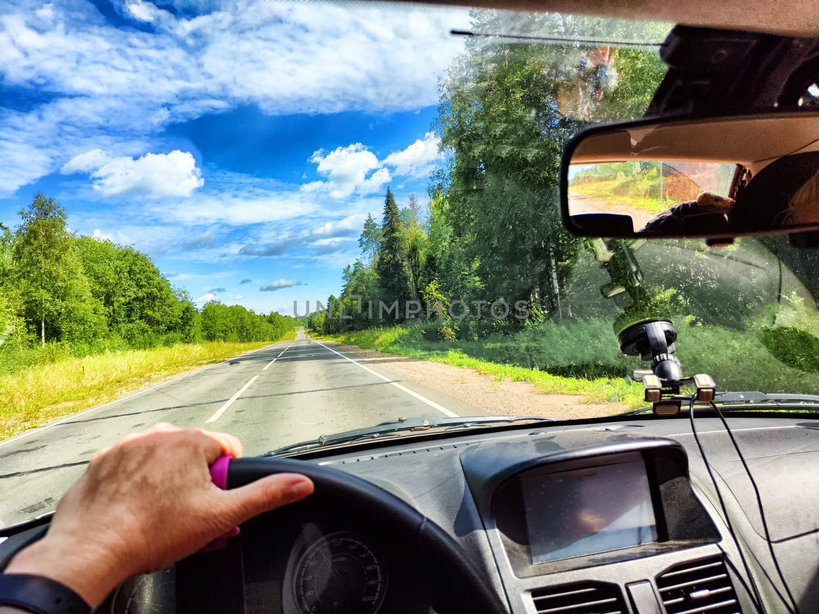 Car salon, steering wheel, hand of woman, view on nature landscape. View from seat of driver on Road, forest, blue sky, white clouds at sunny day. Concept of single trip of female traveller