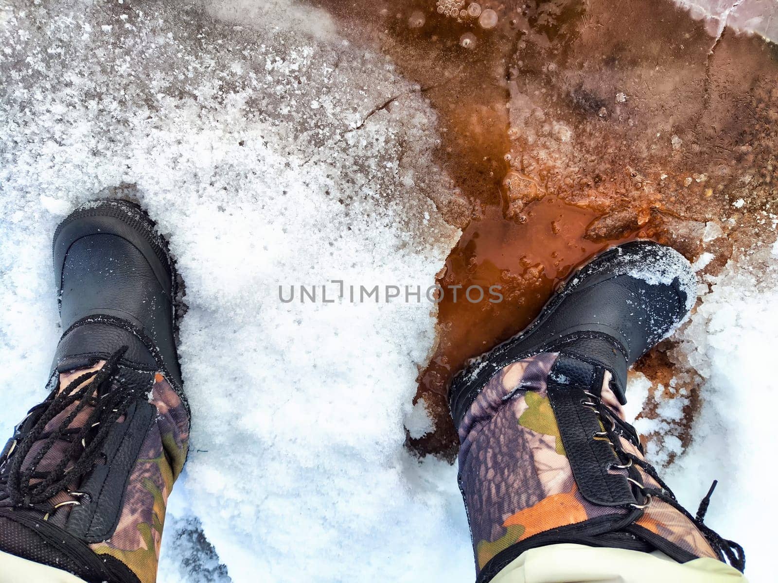 Feet of Hunter or fisherman in big warm boots on winter day on snow. Top view. Fisherman on the ice of a river, lake, reservoir on spring day with melting ice. Dangerous fishing in autumn