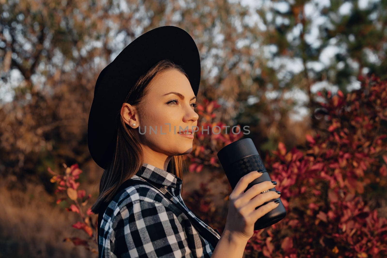 Hipster woman enjoys aroma and taste of fresh coffee. Drinking from thermos in autumn forest. High quality photo