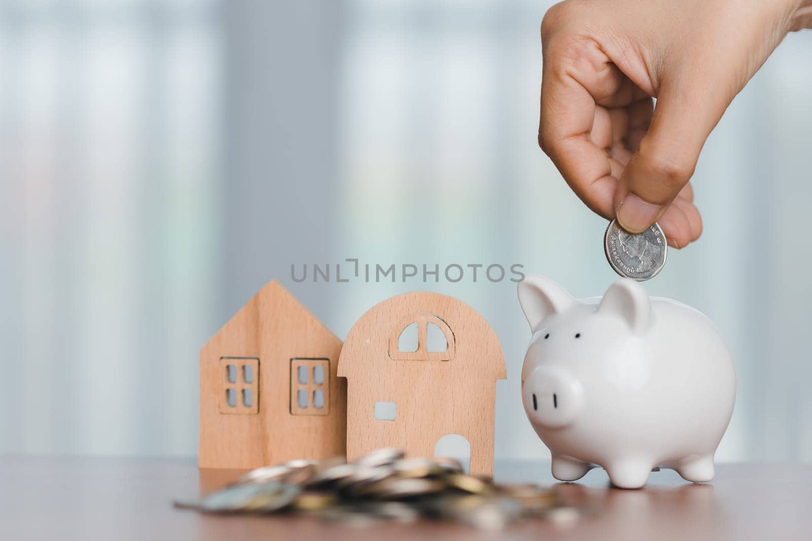 Hand saving money into a piggy bank with the wooden house and a pile of coin for financial growth and home investment concept