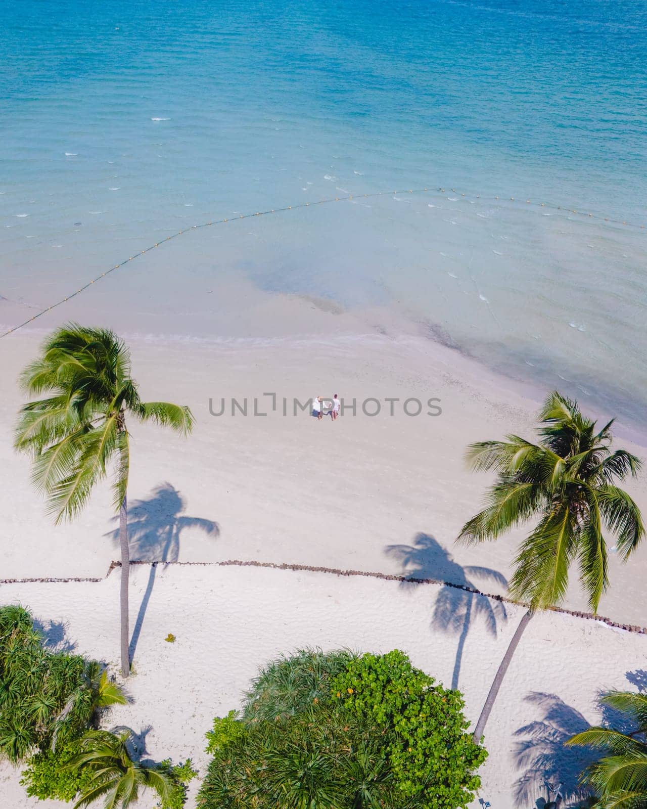 Drone view at a couple walking on the white sandy tropical beach of Koh Muk with palm trees soft white sand, and a turqouse colored ocean in Koh Mook Trang Thailand