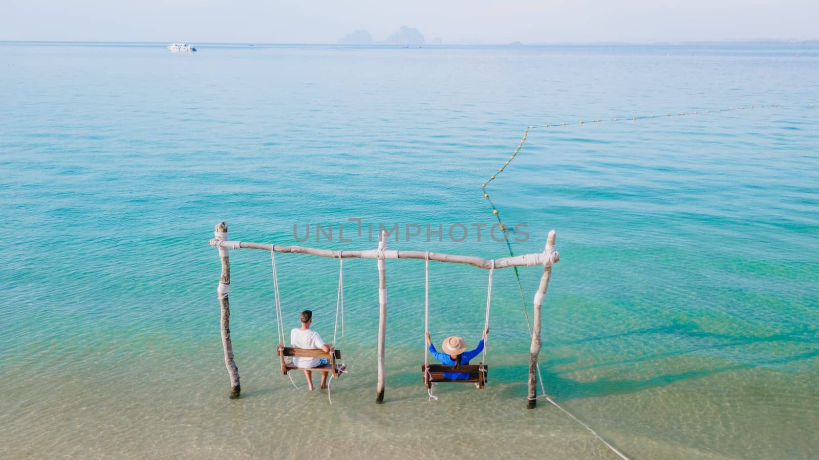 a couple of men and a woman on a swing at the beach of Koh Muk a tropical island with palm trees soft white sand, and a turqouse ocean, Koh Mook Trang Thailand, a couple on a swing in the ocean sunset