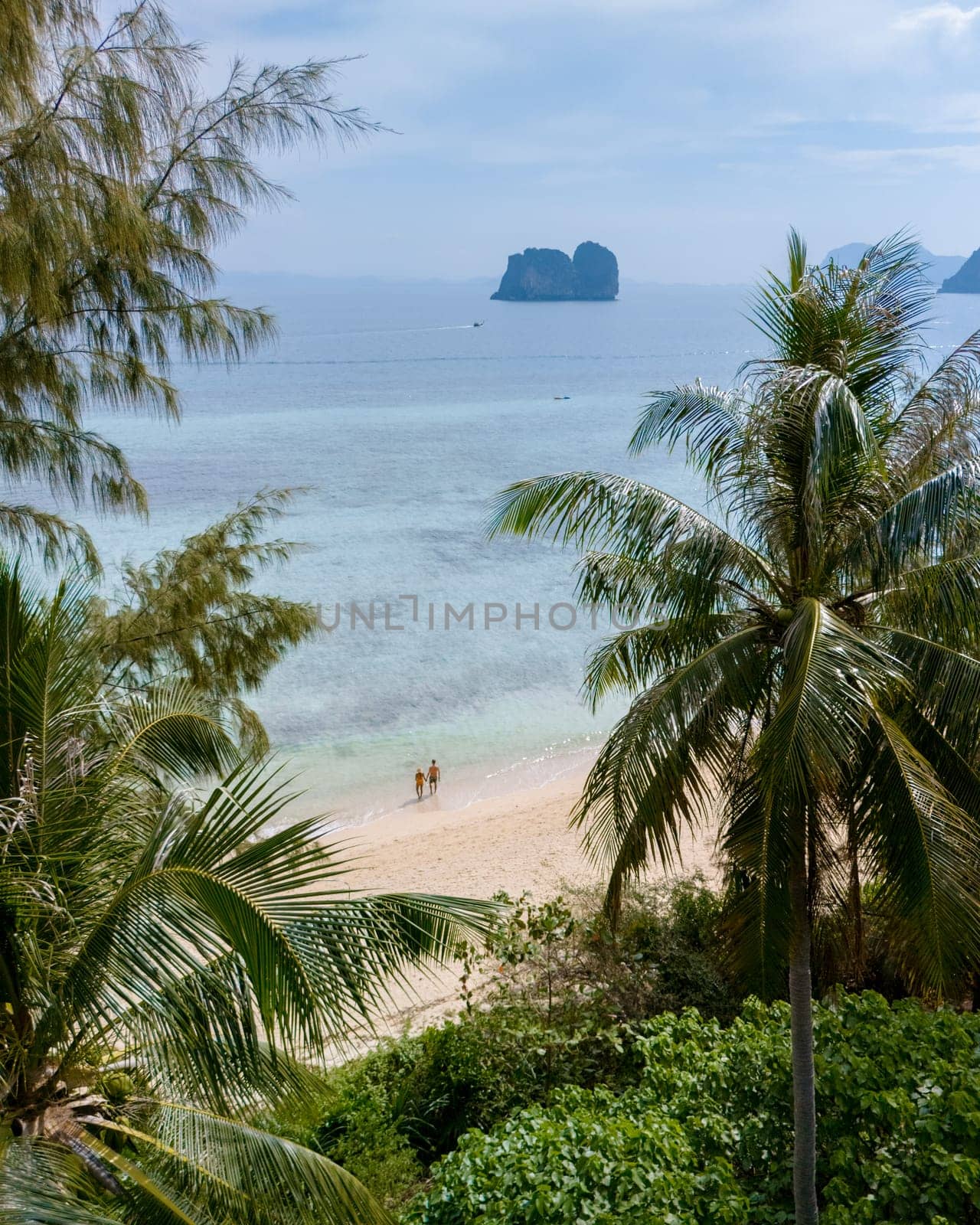 view behind a palm tree at a couple standing at waterfront on the beach at Koh Ngai island, palm trees and soft white sand, and a turqouse colored ocean in Koh Ngai Trang Thailand