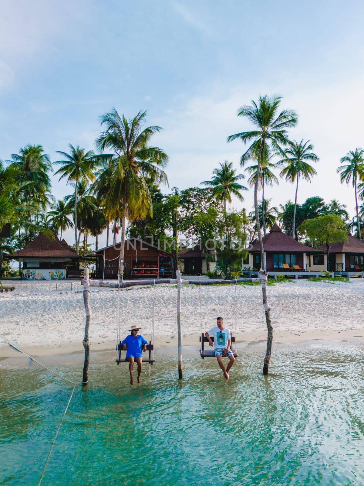 a couple of men and woman on a swing at the beach of Koh Muk a tropical island with palm trees soft white sand, and a turqouse colored ocean in Koh Mook Trang Thailand, couple on a swing in the ocean