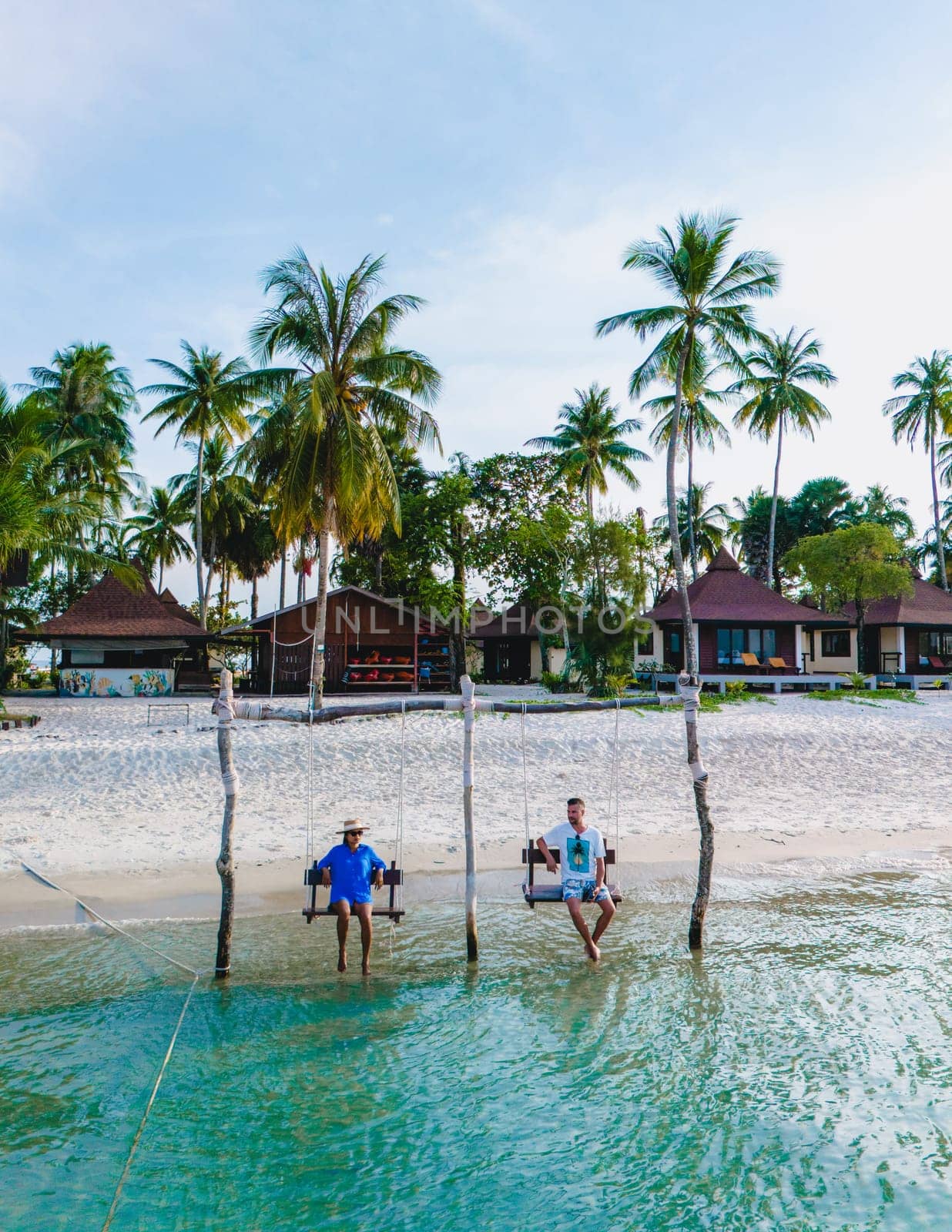 a couple of men and a woman on a swing at the beach of Koh Muk a tropical island with palm trees soft white sand, and a turqouse ocean, Koh Mook Trang Thailand, a couple on a swing in the ocean sunset