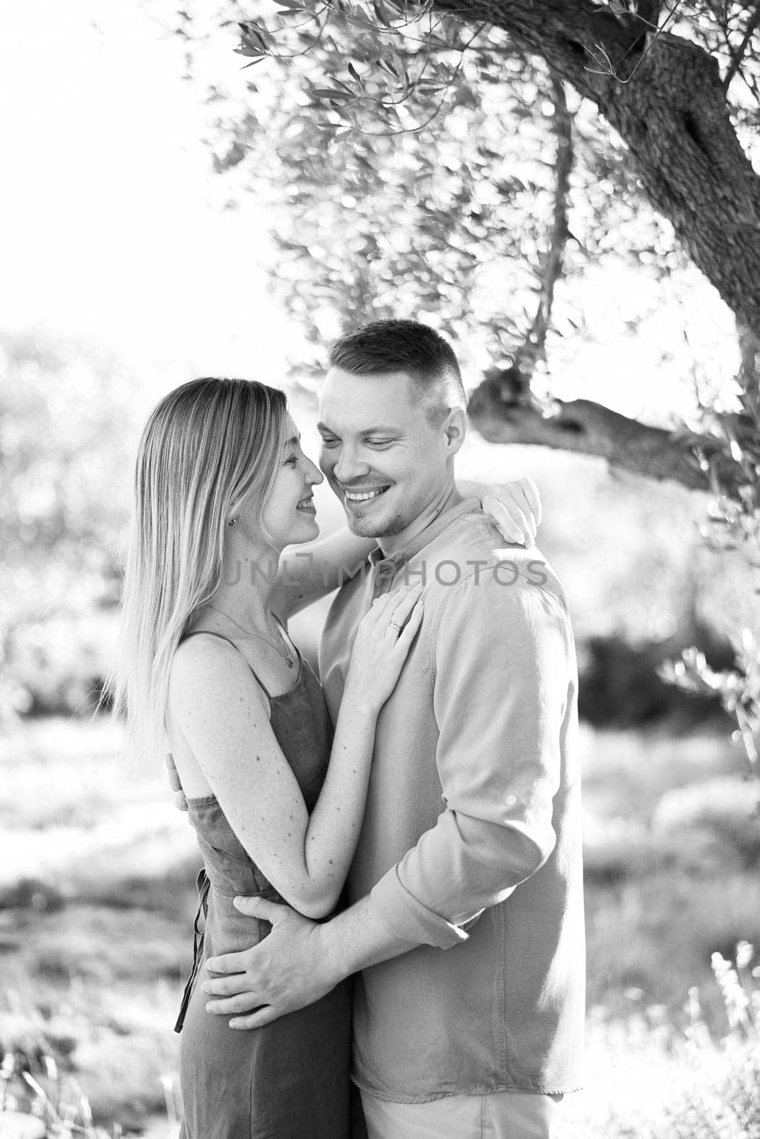 Woman touches her nose to the cheek of smiling man while standing near a tree. Black and white photo by Nadtochiy