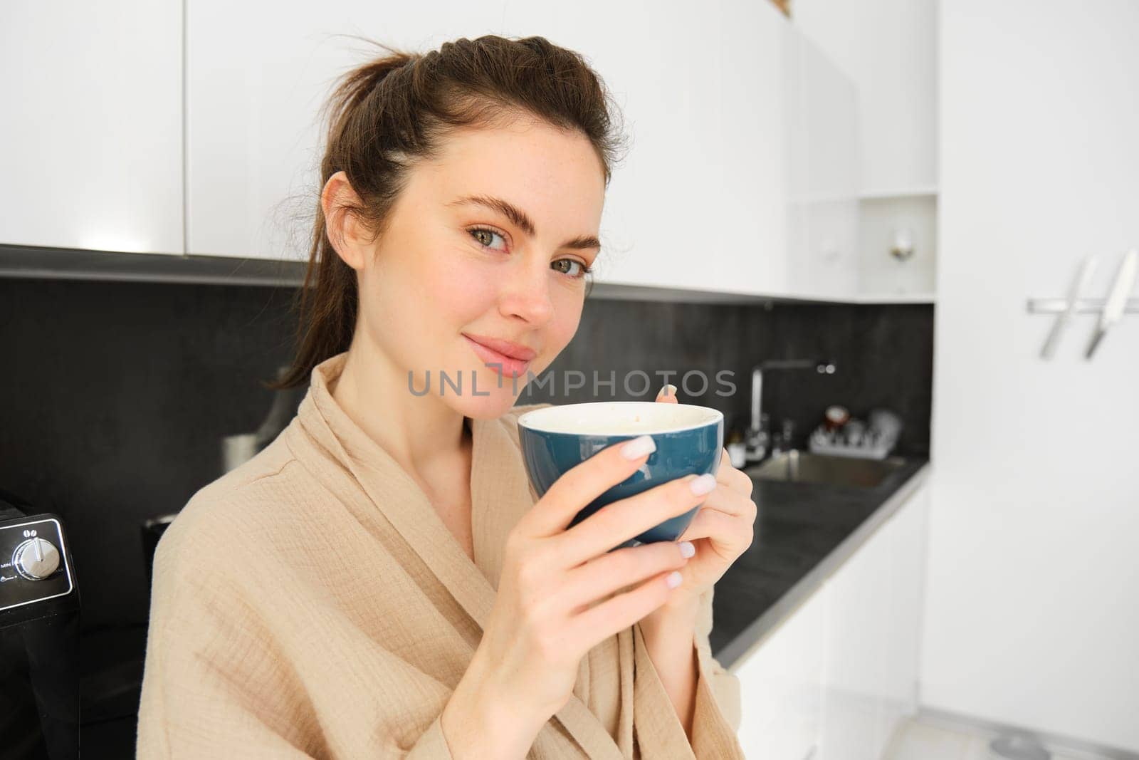 Portrait of beautiful young woman in bathrobe, drinking morning coffee and enjoying the taste, smiling pleased, standing in the kitchen.