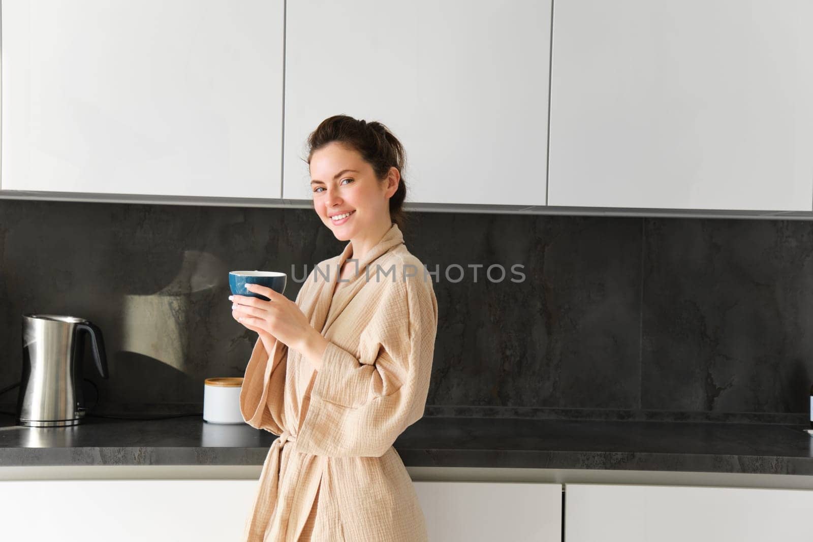 Daily routine and lifestyle. Young beautiful woman in bathrobe, standing in kitchen with cup of coffee, drinking tea, smiling and looking happy.