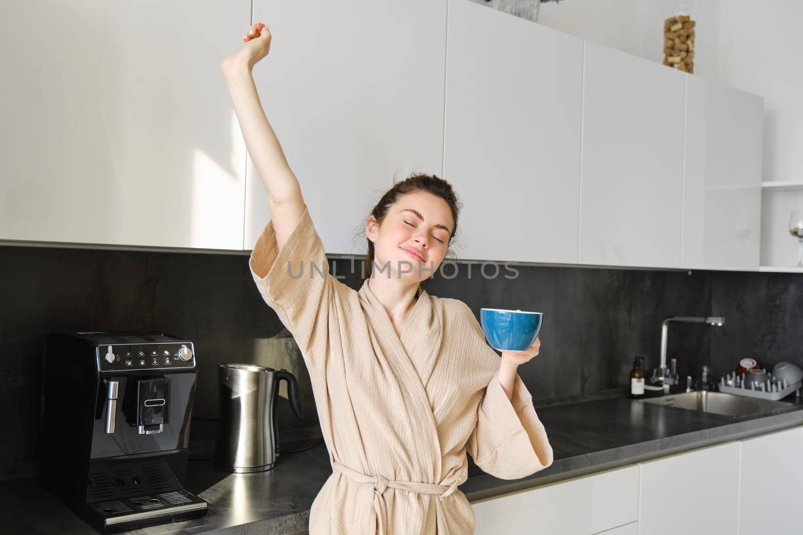 Portrait of carefree brunette woman, dancing and stretching hands in the kitchen, drinking coffee, smiling pleased in the morning.