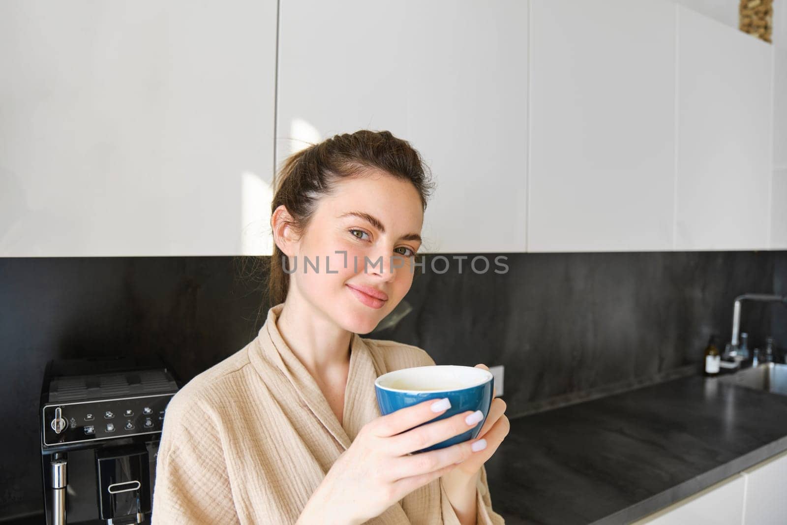 Portrait of good-looking young woman, drinking coffee in the kitchen, enjoying her morning routine and smiling at the camera by Benzoix