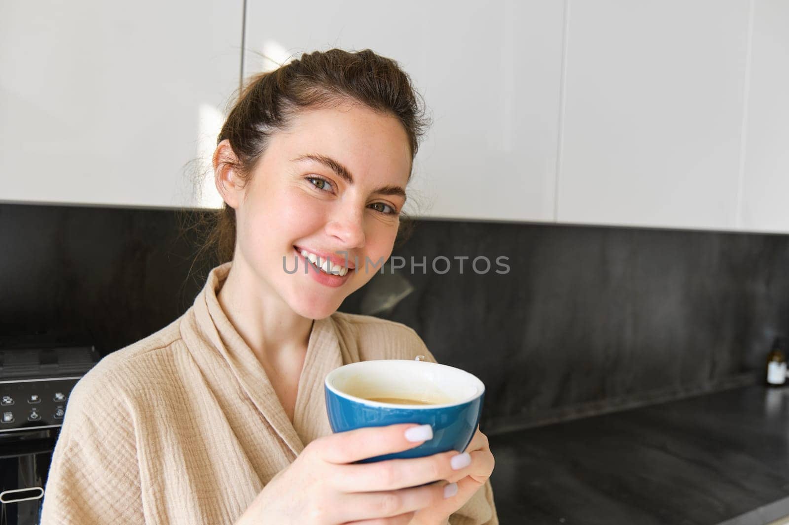 Close up portrait of beautiful, smiling woman, holding cup of coffee, posing in the kitchen, enjoying her morning at home, wearing bathrobe.