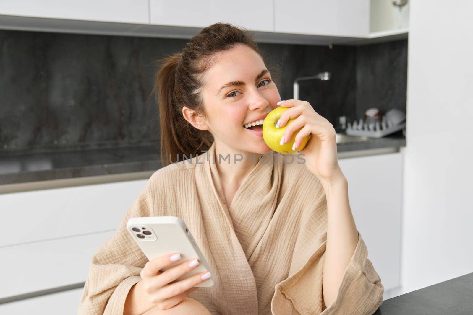 Close up portrait of smiling beautiful woman, sitting in bathrobe, eating apple and looking happy, using smartphone while sitting in the kitchen, order food online on mobile app.