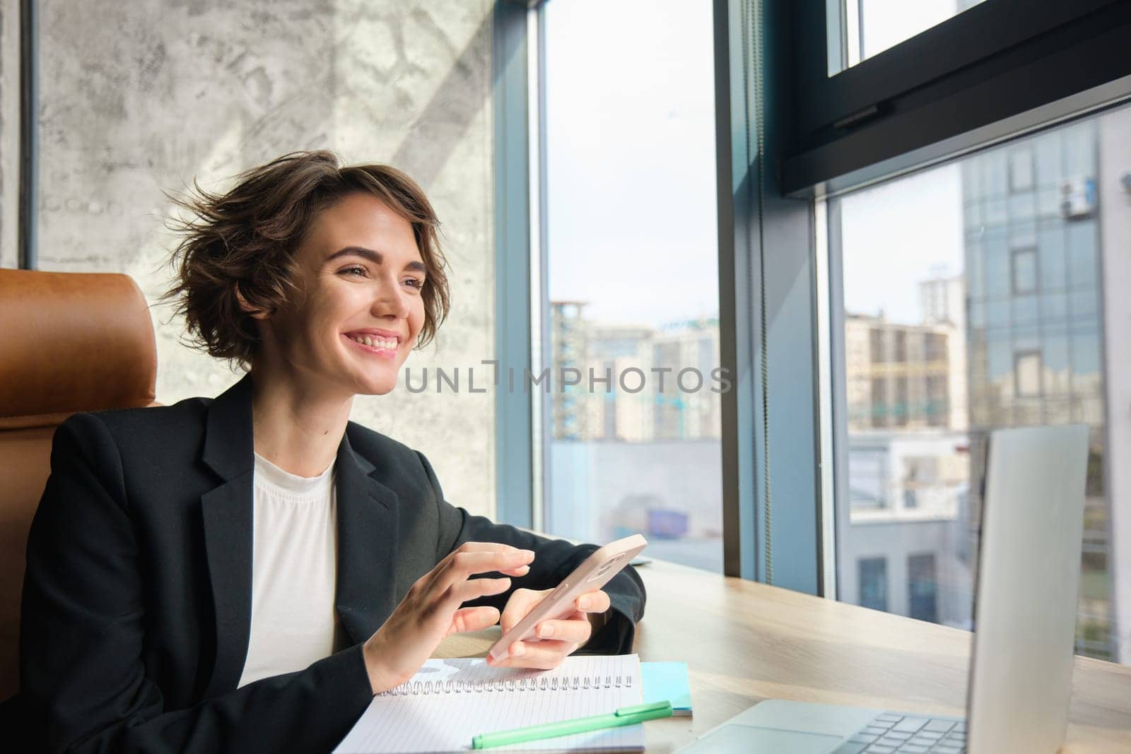 Portrait of businesswoman in suit, sitting in her office in front of window, having a chat on her mobile phone, suing smartphone, has her laptop and documents on table.