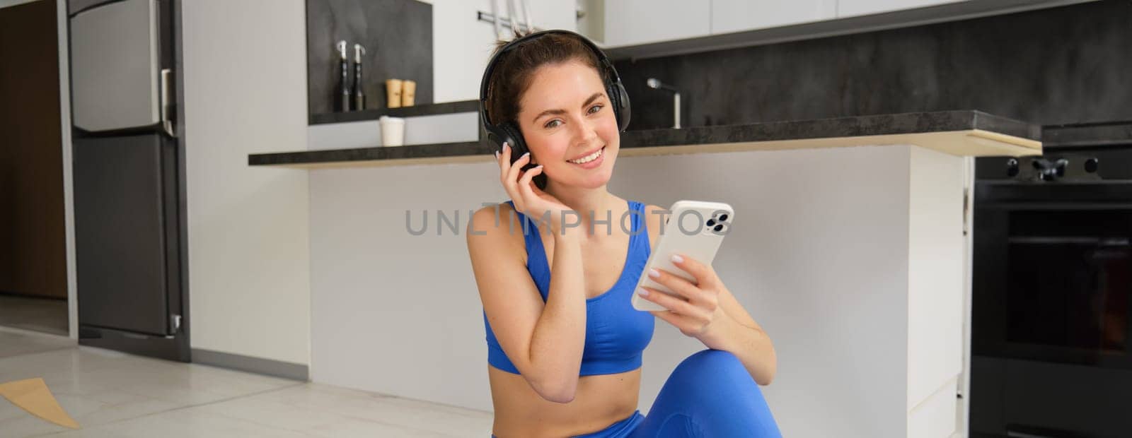 Smiling woman puts on music on smartphone to workout at home, wears wireless headphones, listens to meditation mantra during training session.
