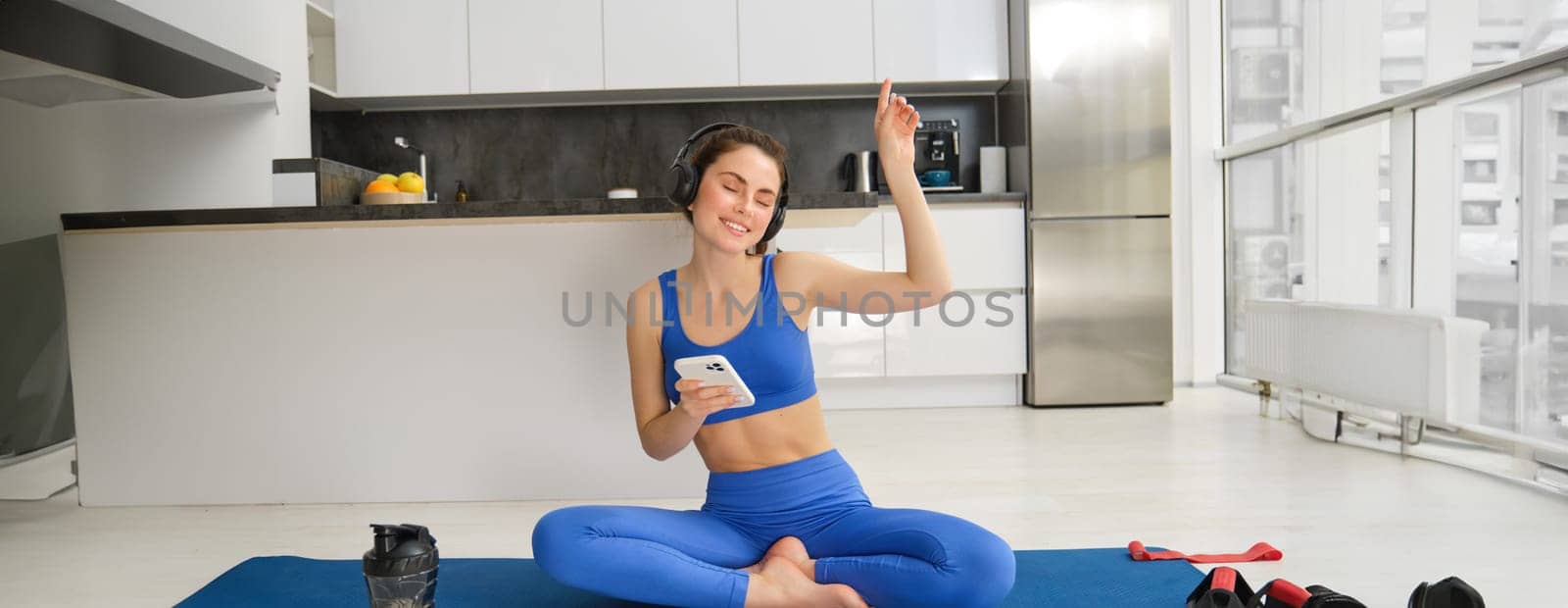 Stylish young fitness instructor, wears activewear, sits on yoga mat and listens music while workout, doing exercises.