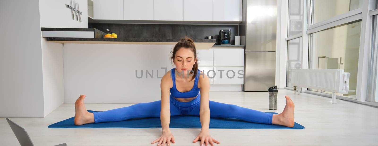 Portrait of young woman does gymnastics training at home, workout, making splits on rubber fitness mat, sitting on floor with feet stretched sideways by Benzoix