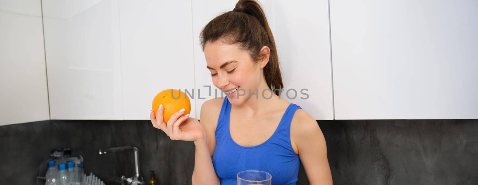 Wellbeing and sport. Young smiling nutritionist, fitness girl holding orange and fresh juice, drinking it from glass and looking happy, posing in sportsbra and leggings, standing in kitchen by Benzoix