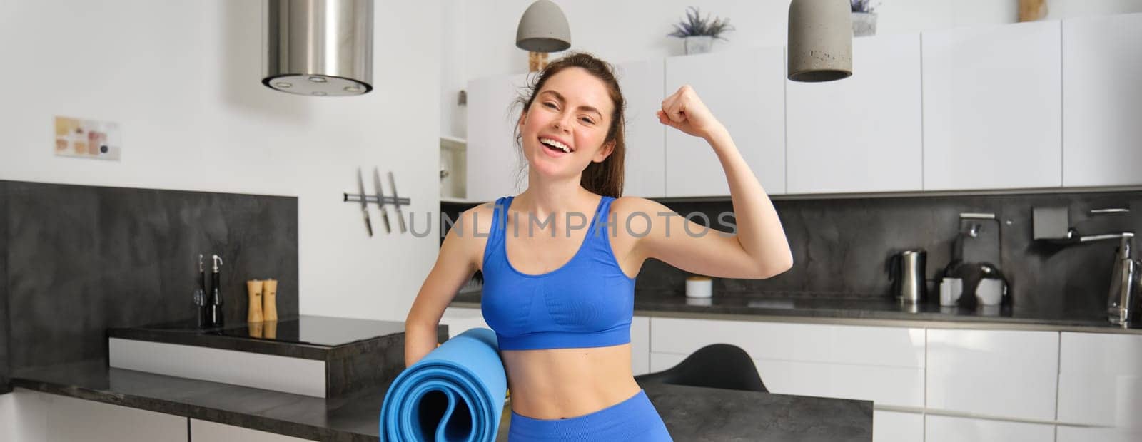 Happy young sportswoman, does fitness at home, holds yoga mat and shows her biceps on arm, flexing her muscles and smiling, standing in living room.
