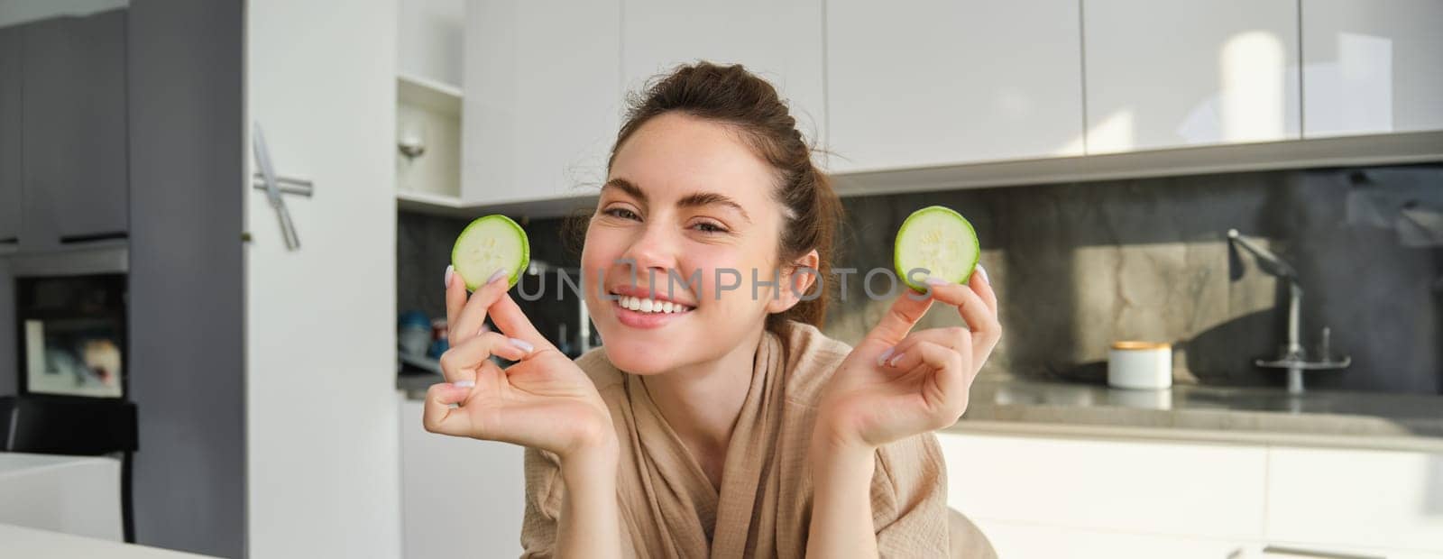 Food and lifestyle concept. Beautiful woman cooking in the kitchen, holding raw zucchini and smiling, preparing healthy vegetarian meal, making salad, looking happy by Benzoix