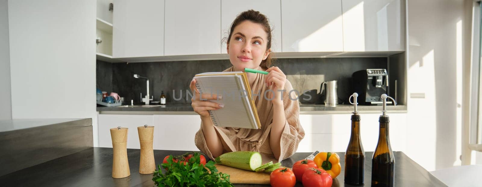 Portrait of woman checking recipe notes in notebook, standing in kitchen with vegetables, cooking food, preparing delicious salad from tomatoes, parsley and olive oil.