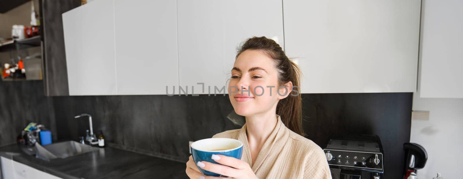 Portrait of smiling woman drinks coffee, stands in the kitchen and enjoys delicious cup of cappuccino in the morning, looks happy at camera by Benzoix