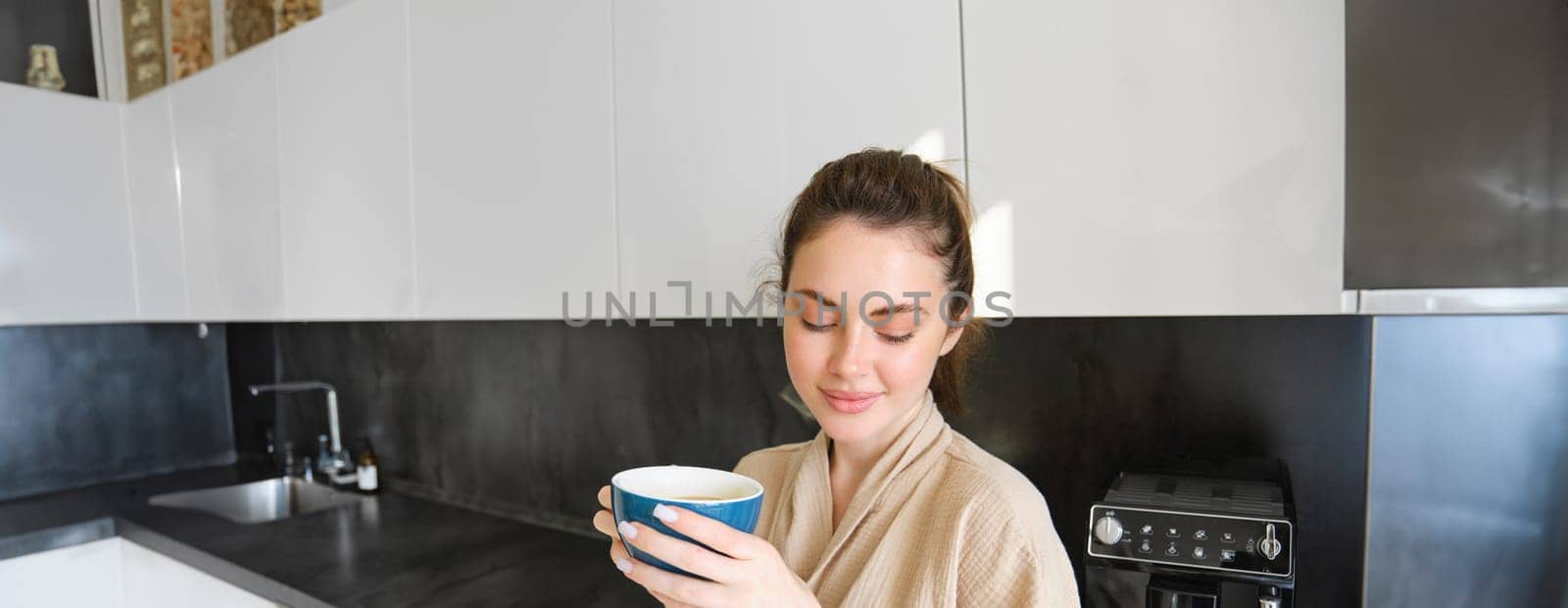 Portrait of smiling woman drinks coffee, stands in the kitchen and enjoys delicious cup of cappuccino in the morning, looks happy at camera.
