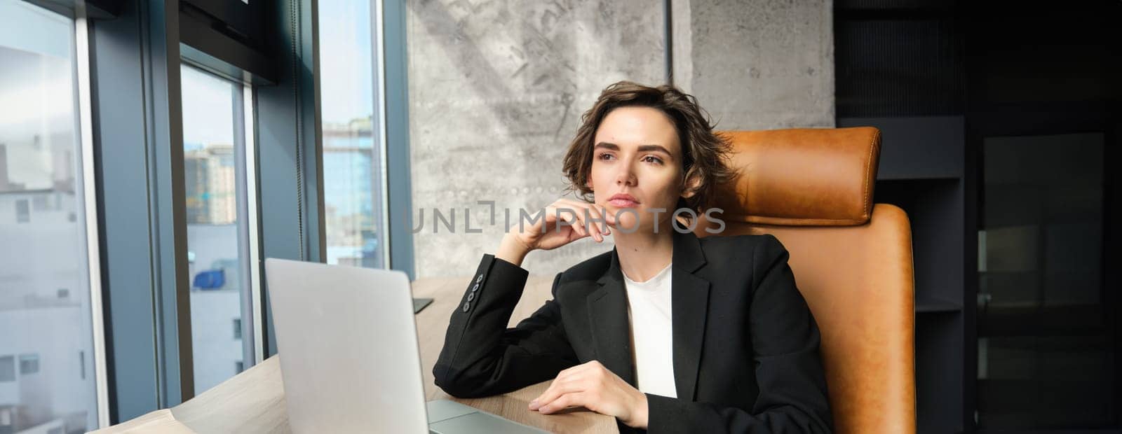 Portrait of confident businesswoman in office, sitting with laptop, looking serious outside window, thinking, solving issue at work.