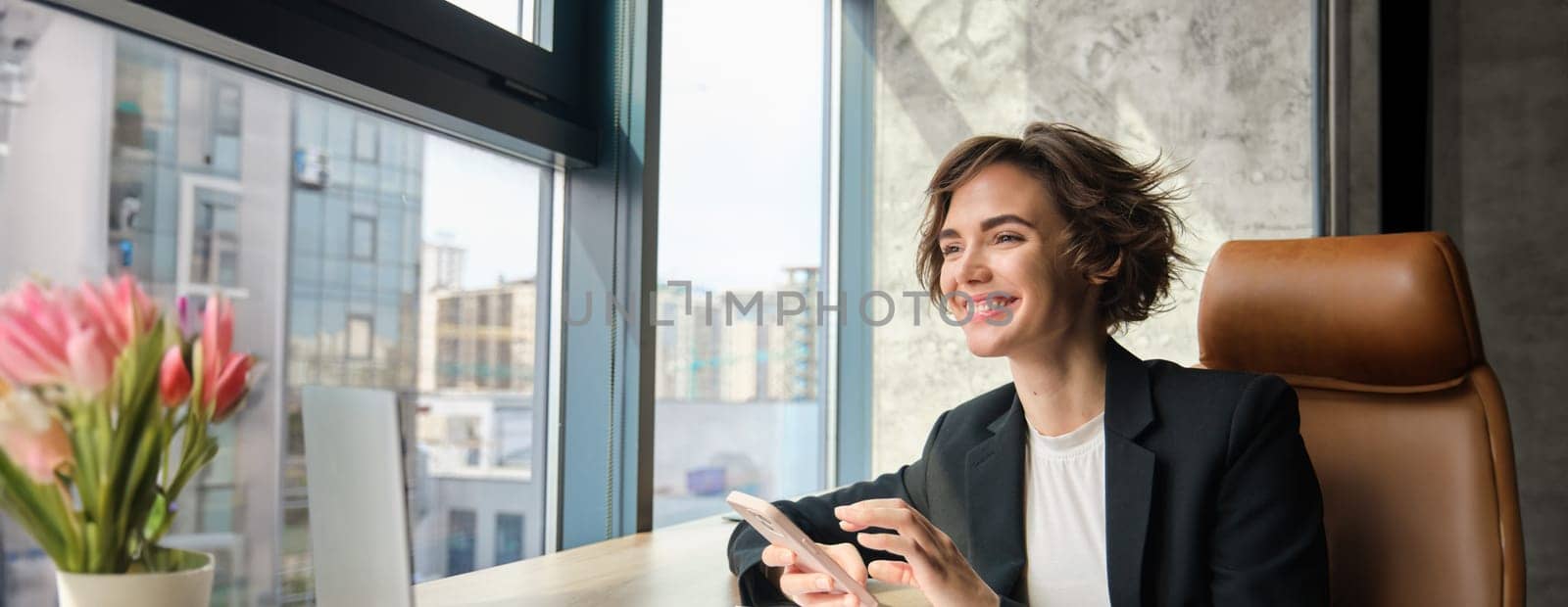 Portrait of businesswoman in suit, sitting in her office in front of window, having a chat on her mobile phone, suing smartphone, has her laptop and documents on table.