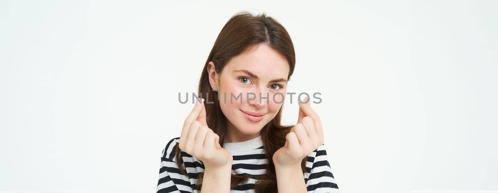 Portrait of cute young brunette woman, shows kawaii finger hearts and smiling, likes smth, stands in striped t-shirt over white background by Benzoix