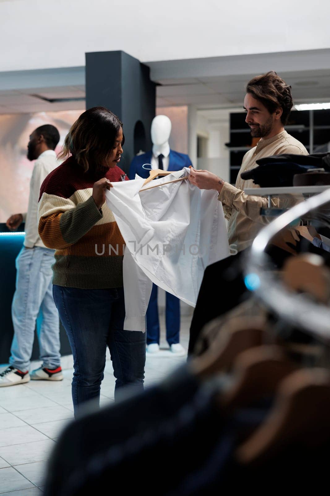 Fashion boutique diverse customer and assistant examining shirt fabric quality together. Clothing store caucasian man consultant providing woman client guidance on size and style