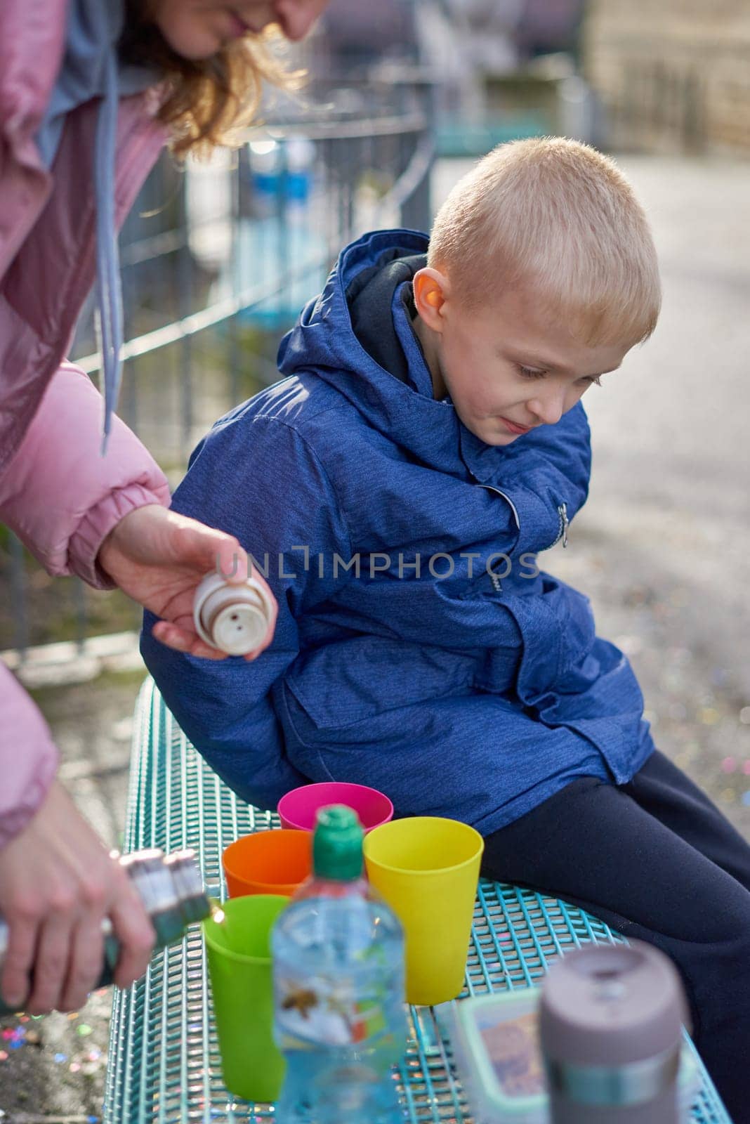 Family Picnic Delight: Cheerful 8-Year-Old Blond Boy in Blue Winter Jacket Sits on Bench While Mom Pours Tea from Thermos, Autumn or Winter. Immerse yourself in the warmth of family moments with this heartening image featuring a joyful 8-year-old blond boy in a blue winter jacket, sitting on a bench while his mom pours tea from a thermos into colorful plastic cups. The photograph captures the essence of a cozy family picnic in the refreshing outdoors during the autumn or winter season.