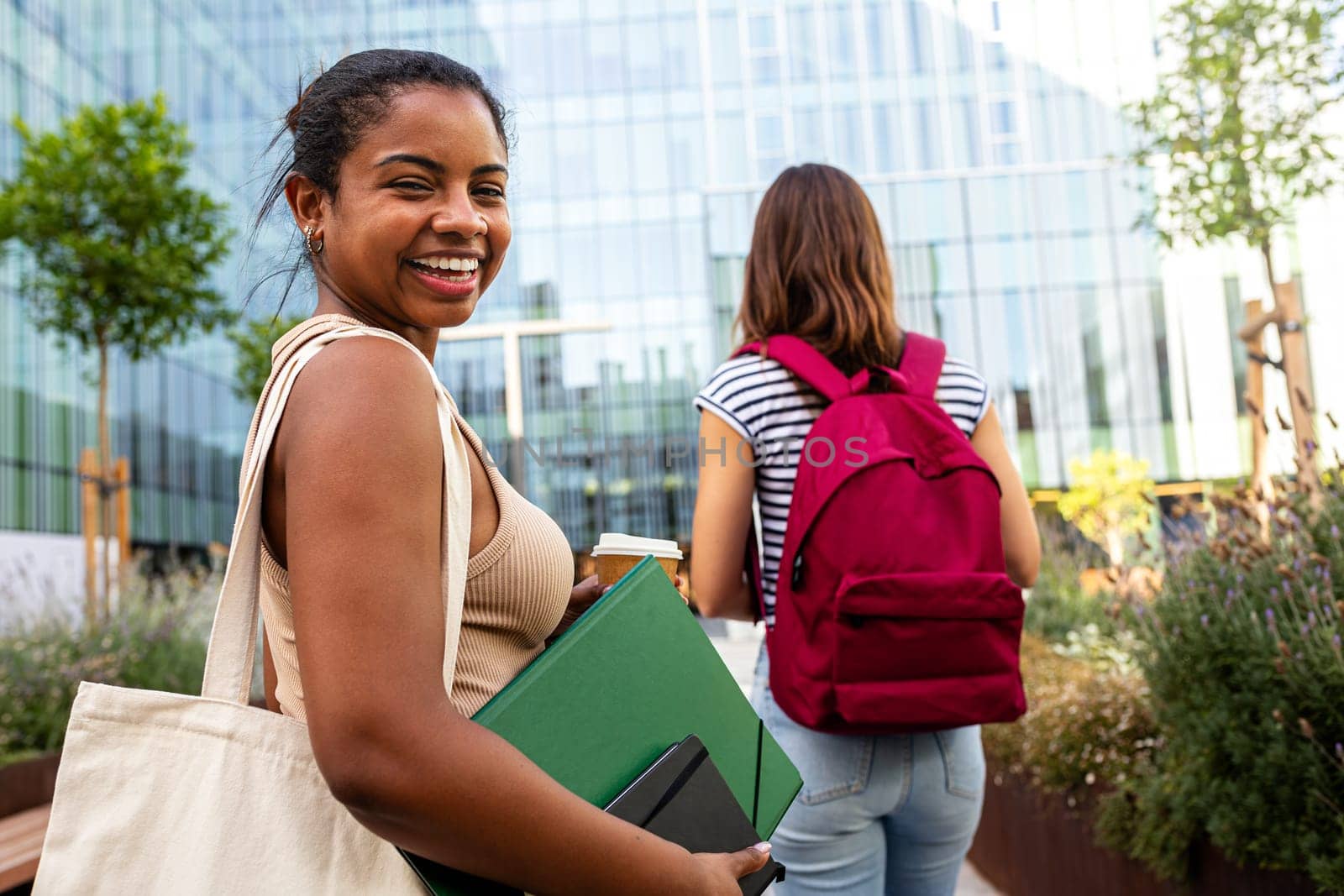 Happy Black female college student standing outside university building holding notebooks in campus looking at camera. by Hoverstock