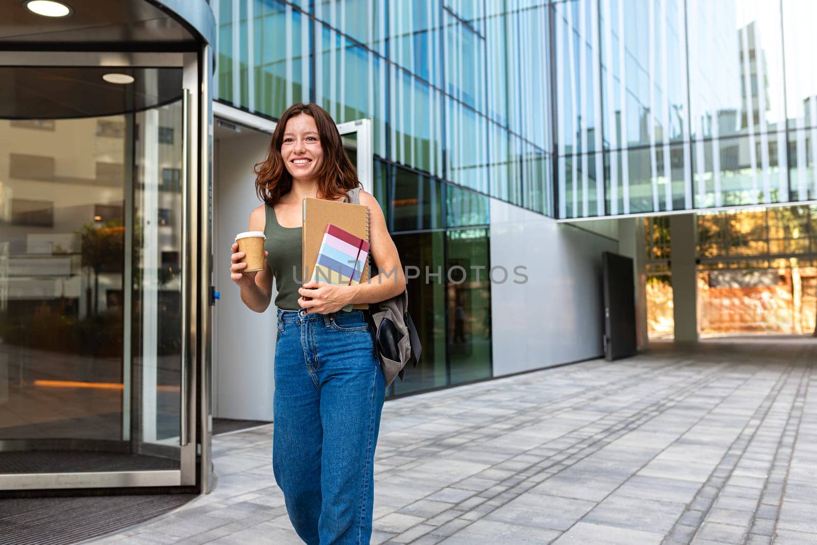 Young redhead happy female university student with backpack walking to class in college campus holding cup of coffee and notebooks. Copy space. Higher education concept.
