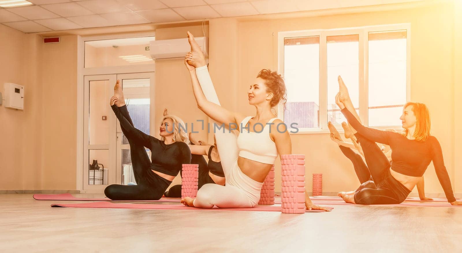 Group of young womans fitness instructor in Sportswear Leggings and Tops, stretching in the gym before pilates, on a yoga mat near the large window on a sunny day, female fitness yoga routine concept.