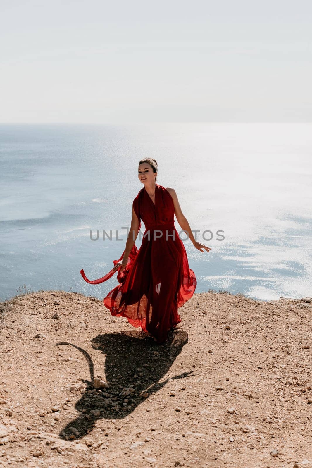 Side view a Young beautiful sensual woman in a red long dress posing on a rock high above the sea during sunrise. Girl on the nature on blue sky background. Fashion photo.