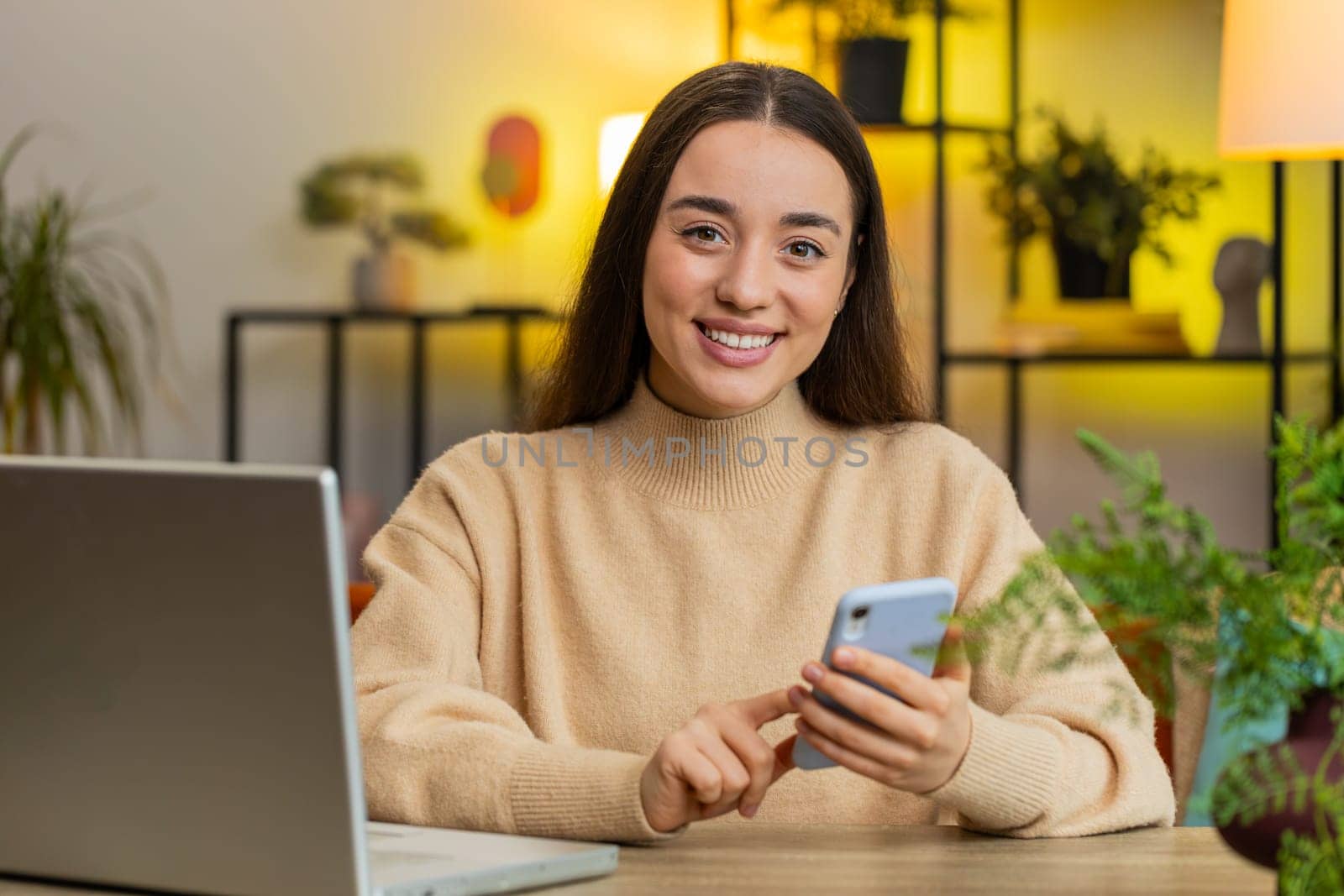 Happy young woman sits at table uses mobile phone smiles at home office room apartment. Girl lady texting share messages content on smartphone social media applications online watching relax movie