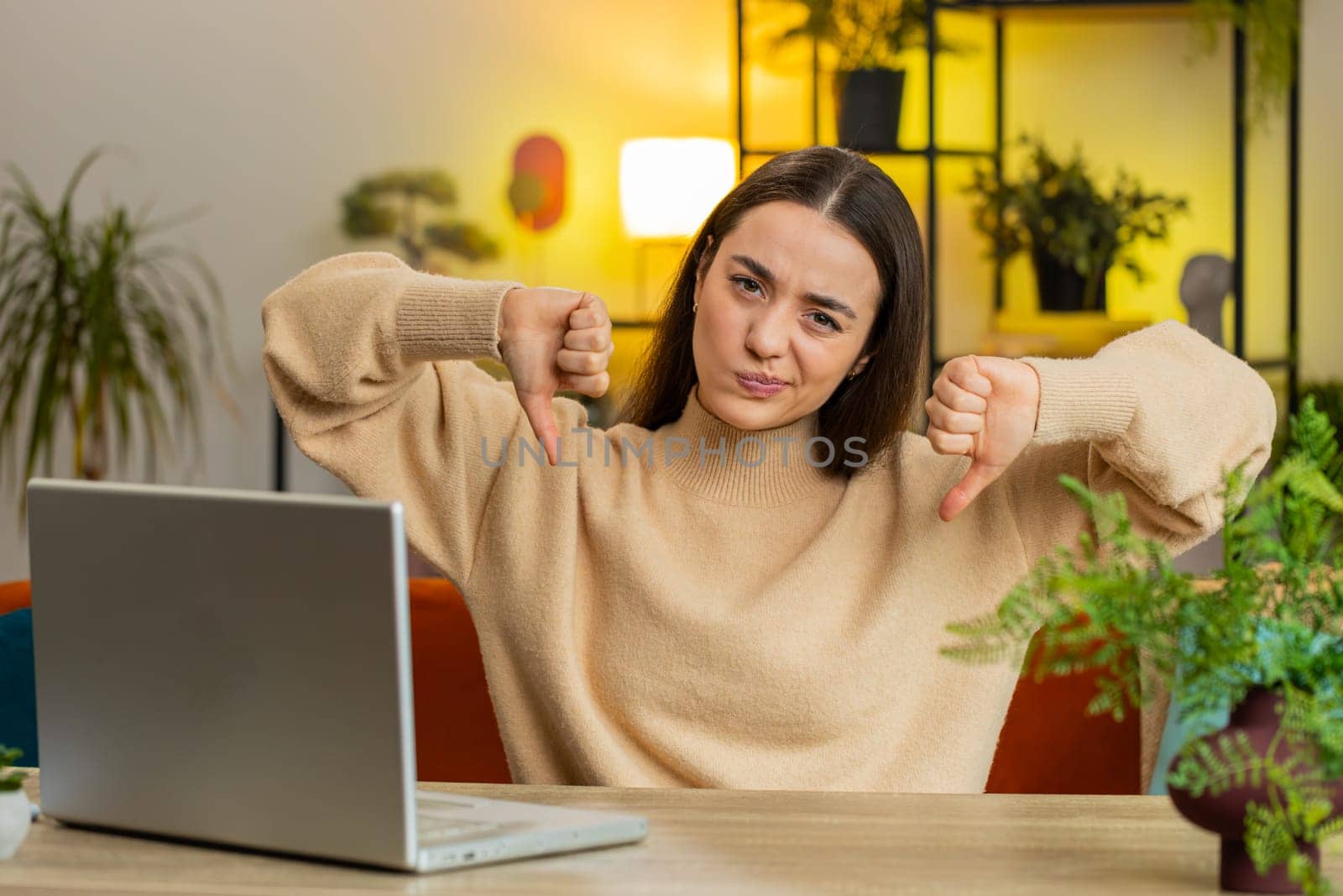 Dislike. Upset unhappy Caucasian woman showing thumbs down hands sign gesture expressing discontent disapproval dissatisfied bad work at home office indoors. Displeased young girl in room at table