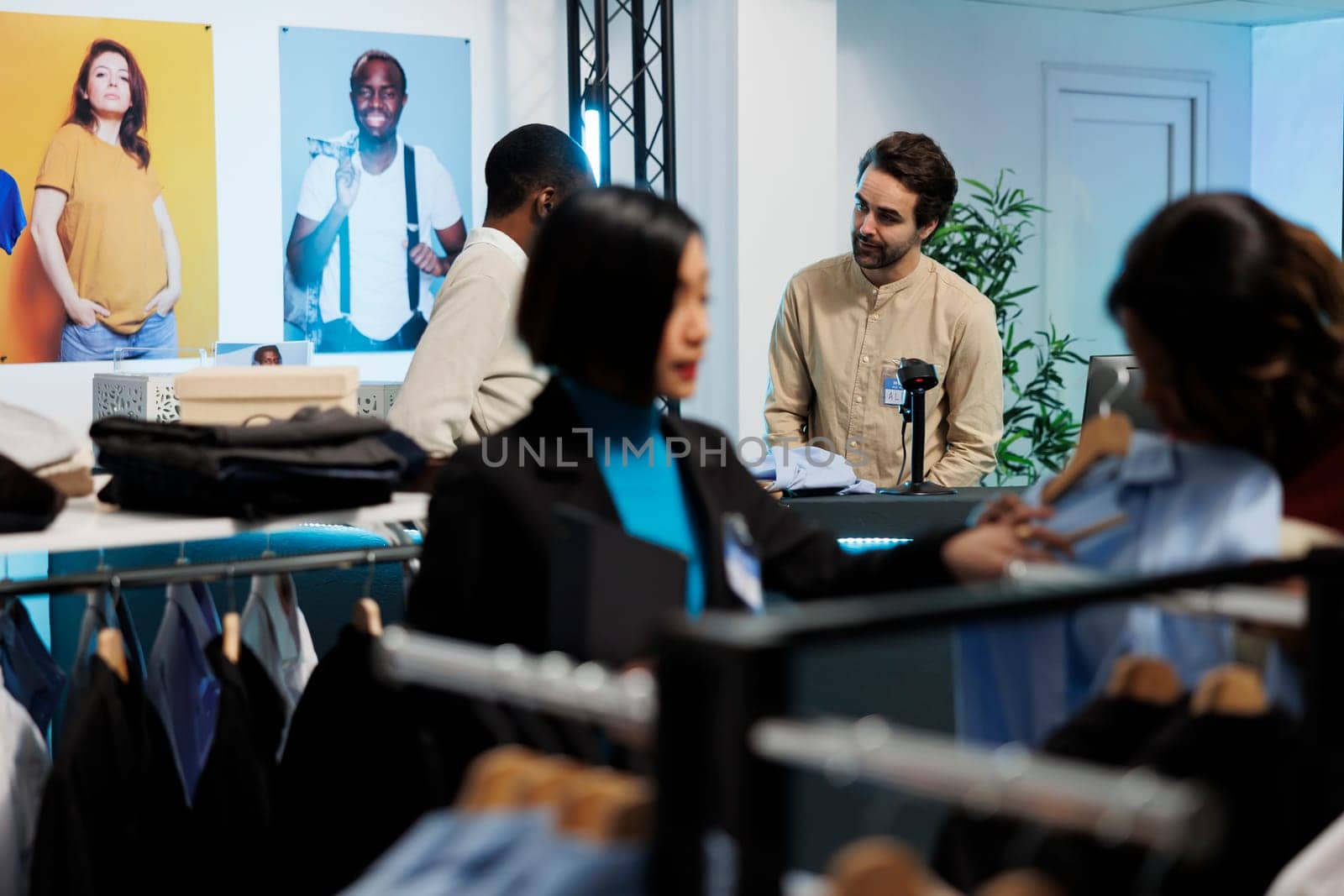 Clothing store cashier at clothing store engaging in conversation with customer at counter. Buyer waiting in line while man employee scanning apparel and talking with client