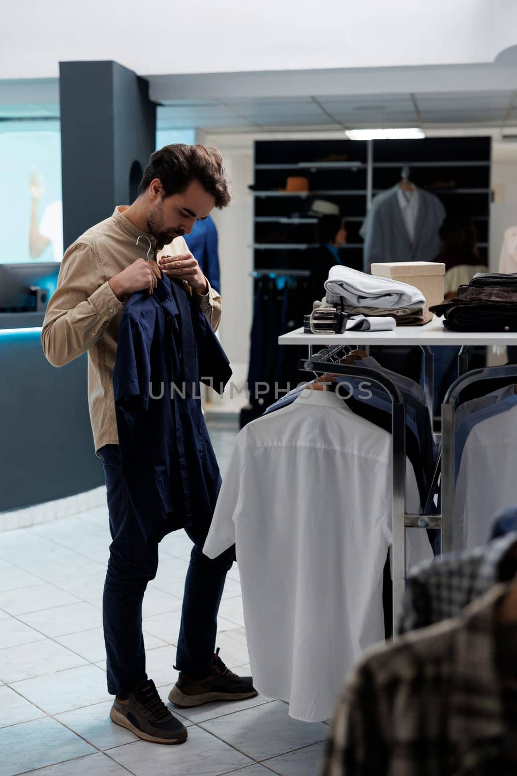 Customer holding shirt to body while choosing formal outfit in fashion boutique menswear department. Caucasian man trying on trendy apparel and examining style before buying