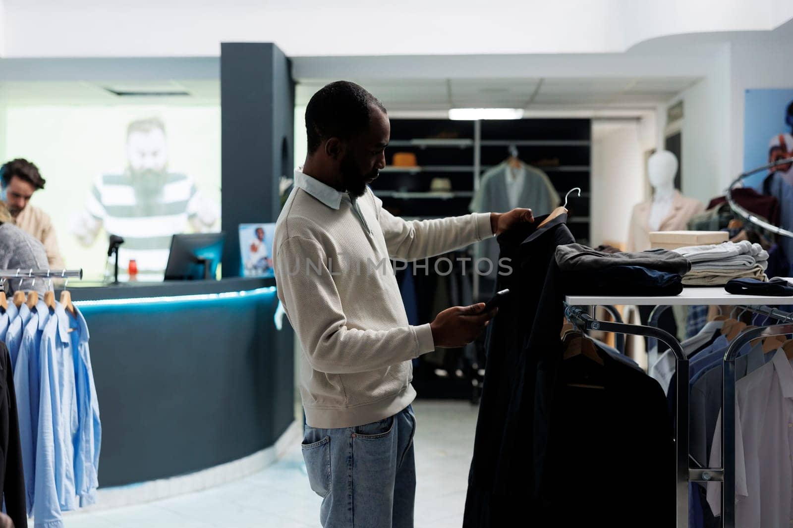 Clothing store customer holding apparel on hanger and checking available size in stock on mobile phone site. Man examining garment and scrolling boutique smartphone application