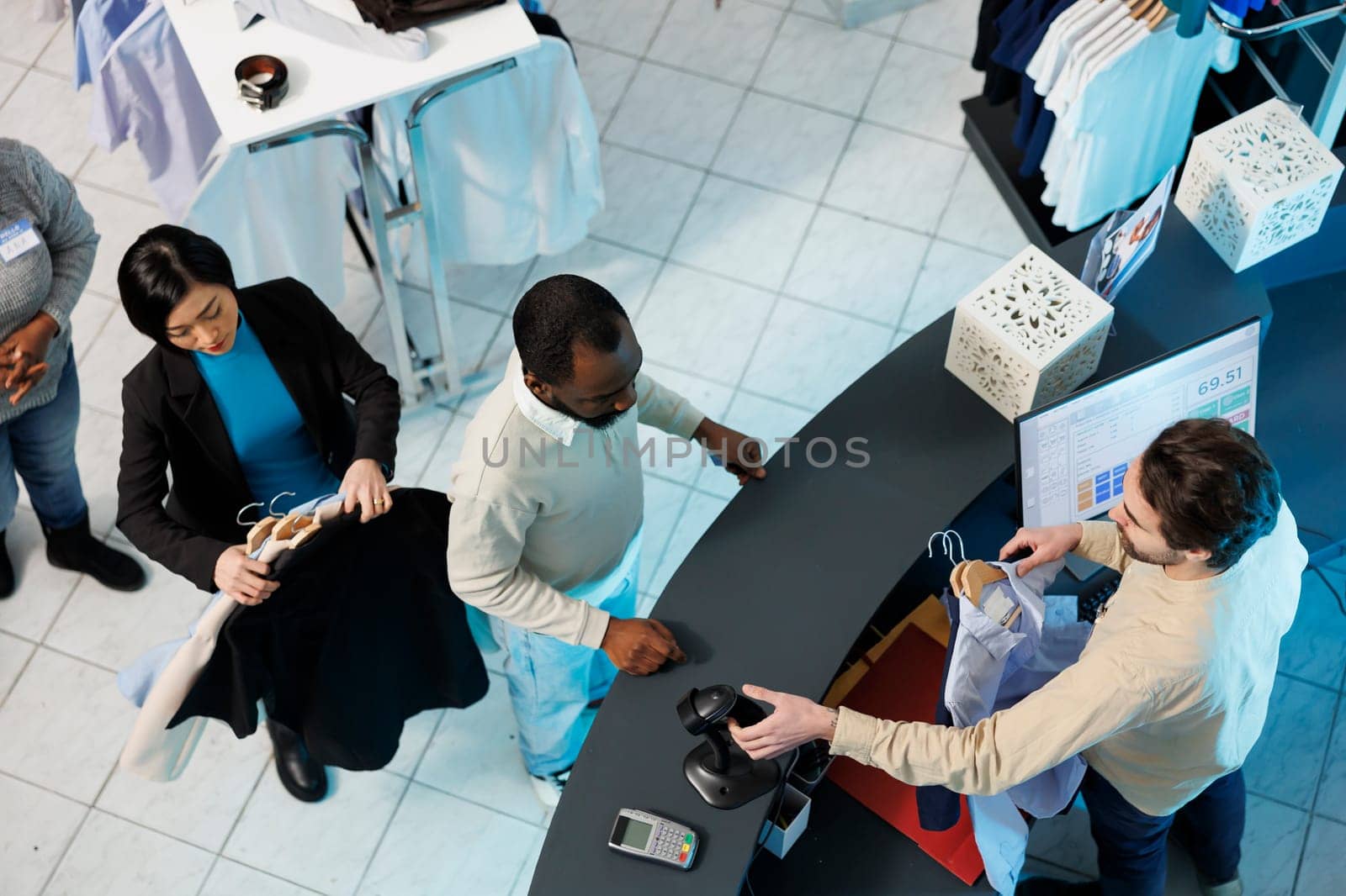 Cashier at clothing store checkout desk scanning and processing purchase for customer top view. Diverse people standing in line at counter desk while boutique employee using scanner