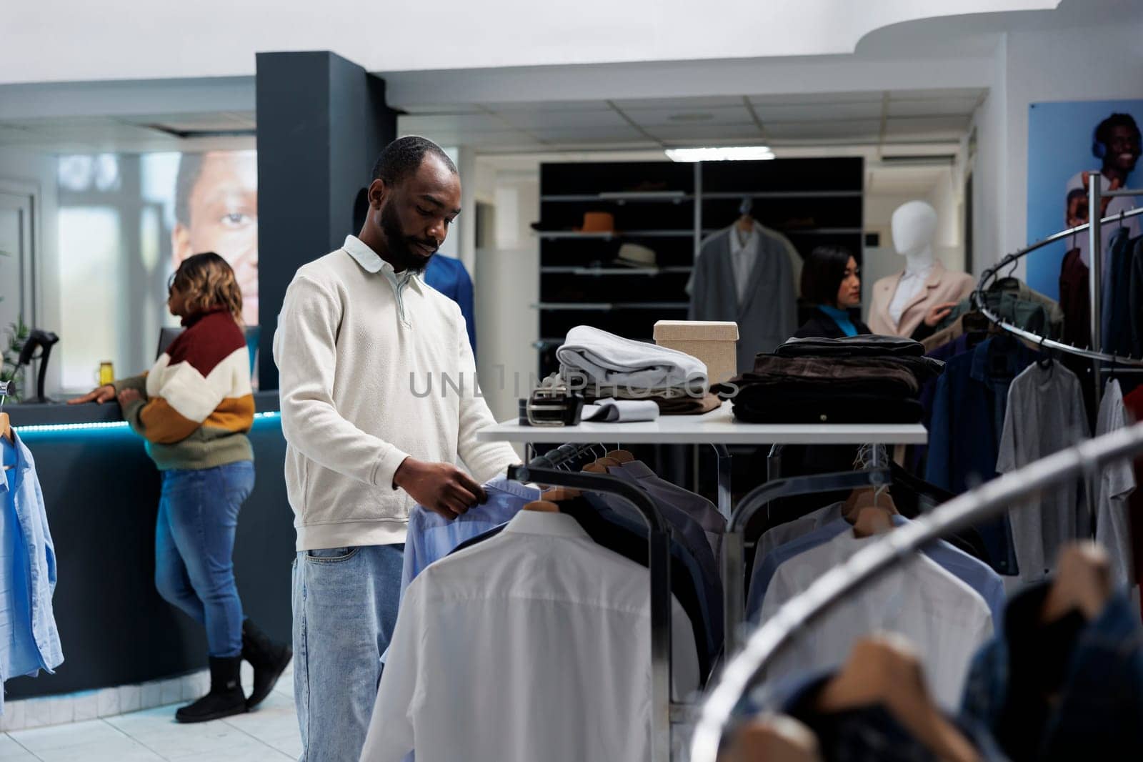 Young african american man shopping for casual apparel and browsing rack with shirts in menswear boutique. Clothing store customer choosing outfit and checking garment style