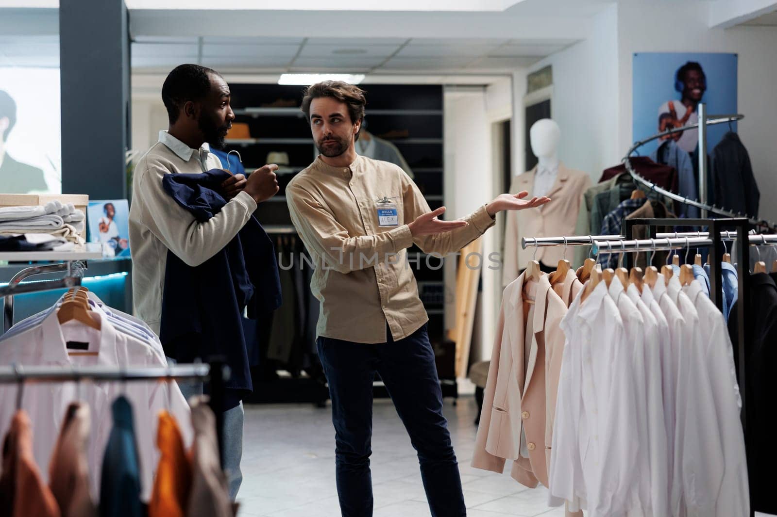 Shopping mall worker pointing at apparel rack while offering customer help in selecting casual outfit. Clothing store consultant assisting african american man in choosing garment