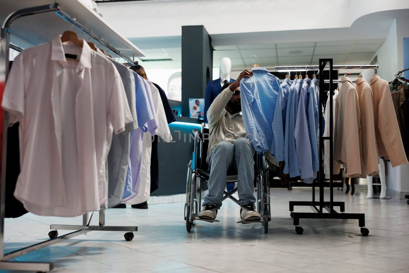 African american man in wheelchair browsing through rack and choosing casual shirt in clothing shop. Shopping center boutique customer with physical disability examining apparel