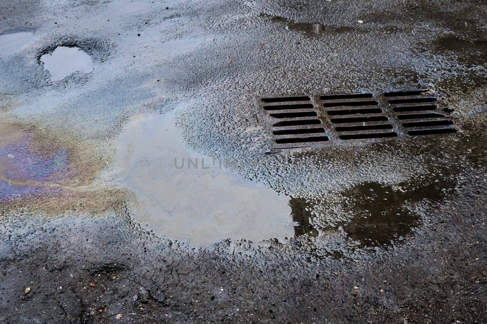 Oil or gasoline stains caught in a puddle against the background of an old asphalt surface with a grate flowing into a storm drain. Environmental problems of water pollution.