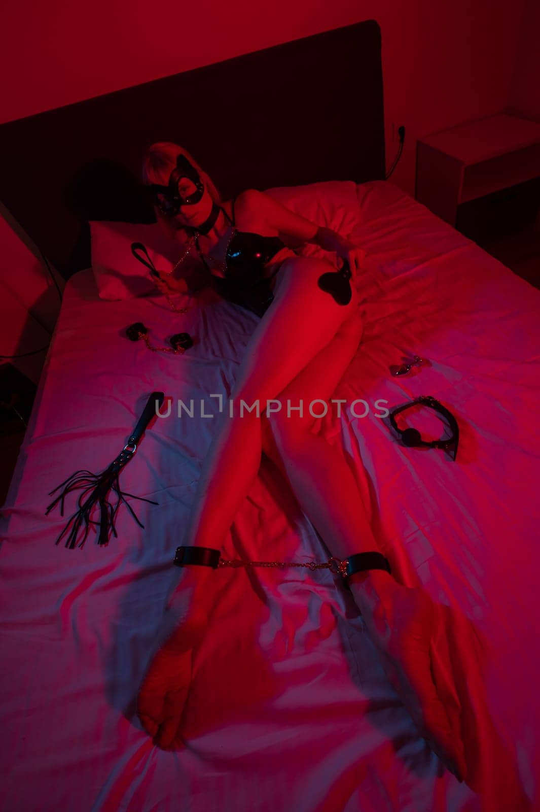 A woman in chains on her legs lies on a bed in a neon red-blue light. BDSM concept