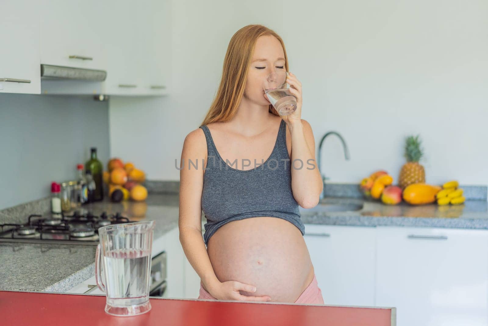 Embracing the vital benefits of water during pregnancy, a pregnant woman stands in the kitchen with a glass, highlighting hydration's crucial role in maternal well-being by galitskaya