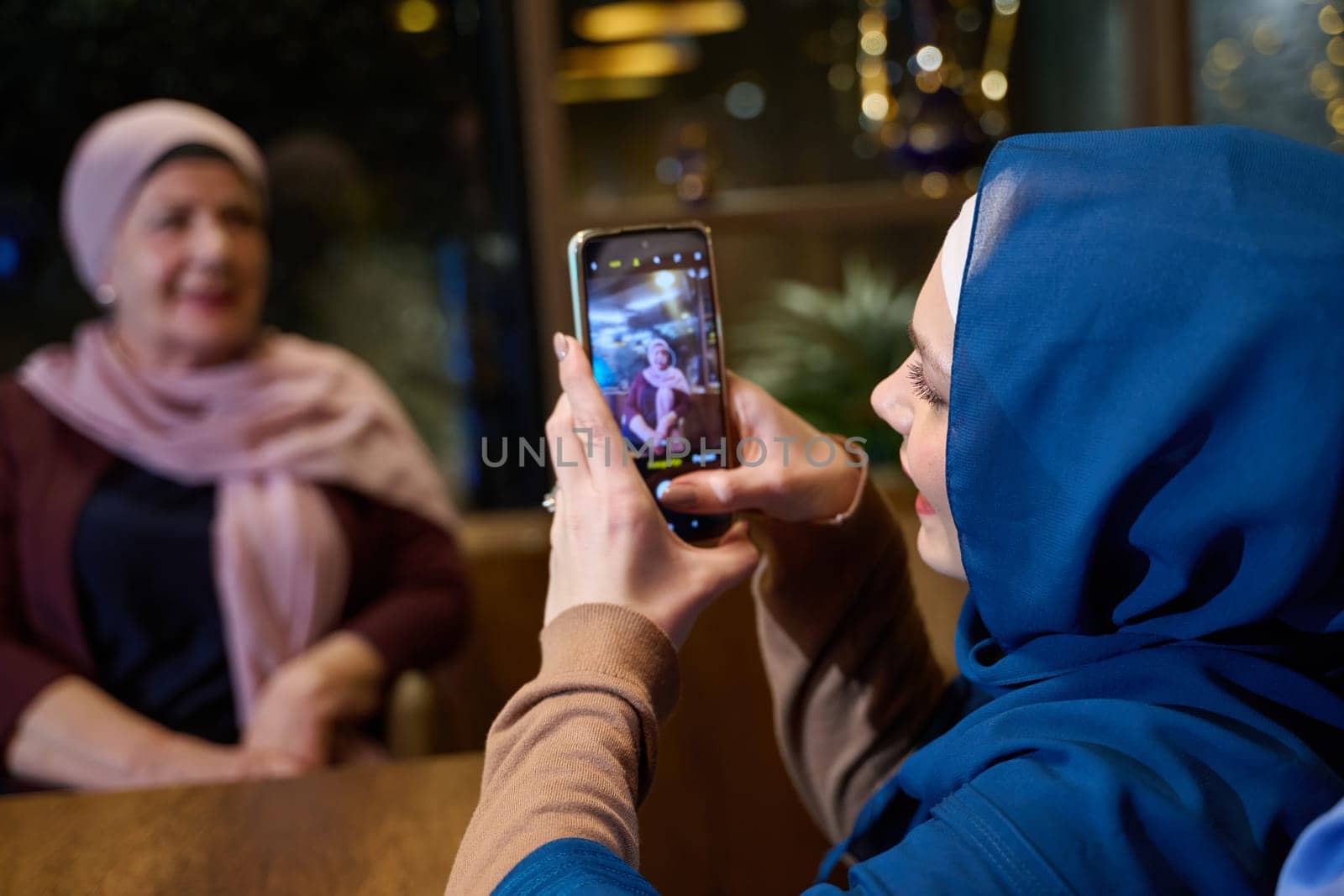 In a modern restaurant during the holy month of Ramadan, a woman in a hijab captures a moment with her mother using a smartphone, epitomizing the blending of tradition and technology in familial bonds and capturing cherished memories.