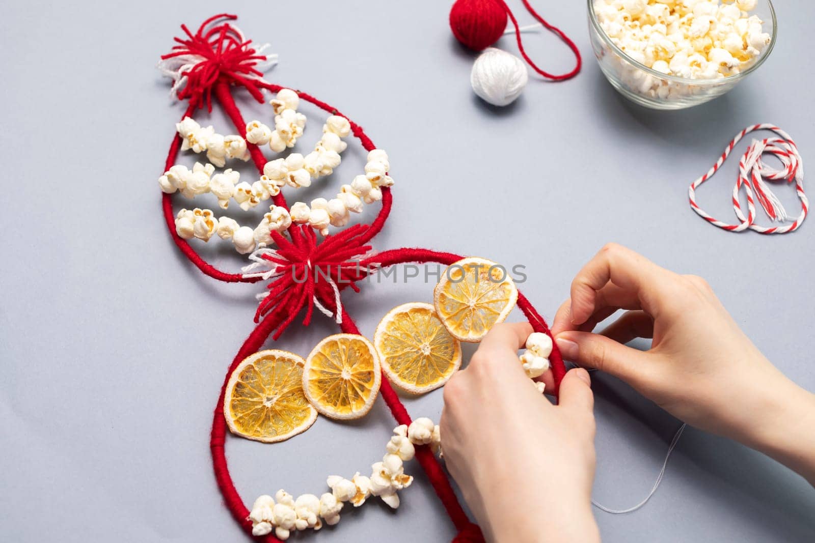 Female hands make Survakane, Survaknitsa, Survachka, Surva. Bulgarian Traditional Christmas Cornel Tree Twig. Flat lay with Popcorn, Red White Threads, Dried Orange. World Tree. Horizontal plane.