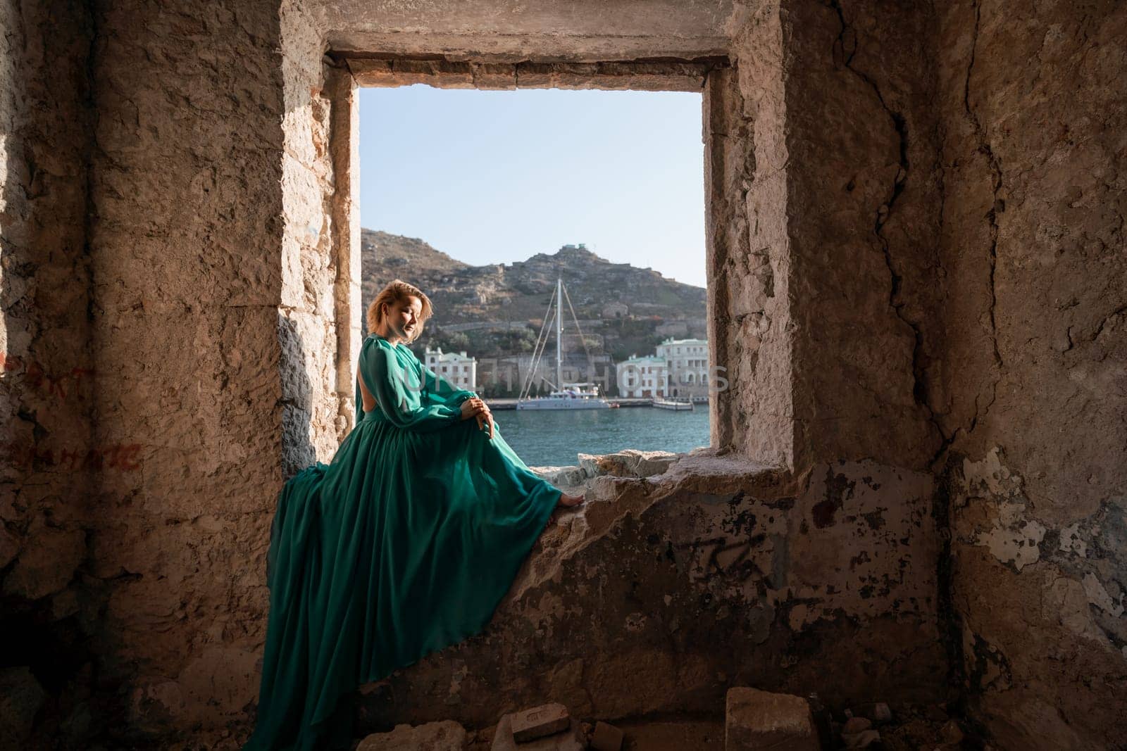 Rear view of a happy blonde woman in a long mint dress posing against the backdrop of the sea in an old building with columns. Girl in nature against the blue sky. by Matiunina